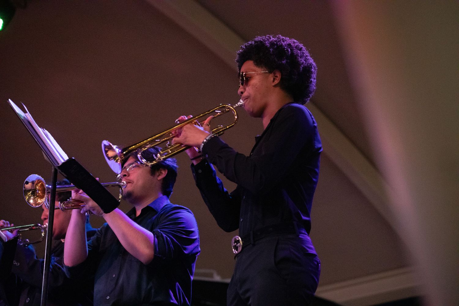 Trumpet players perform in silhouette during a concert with purple stage lighting.