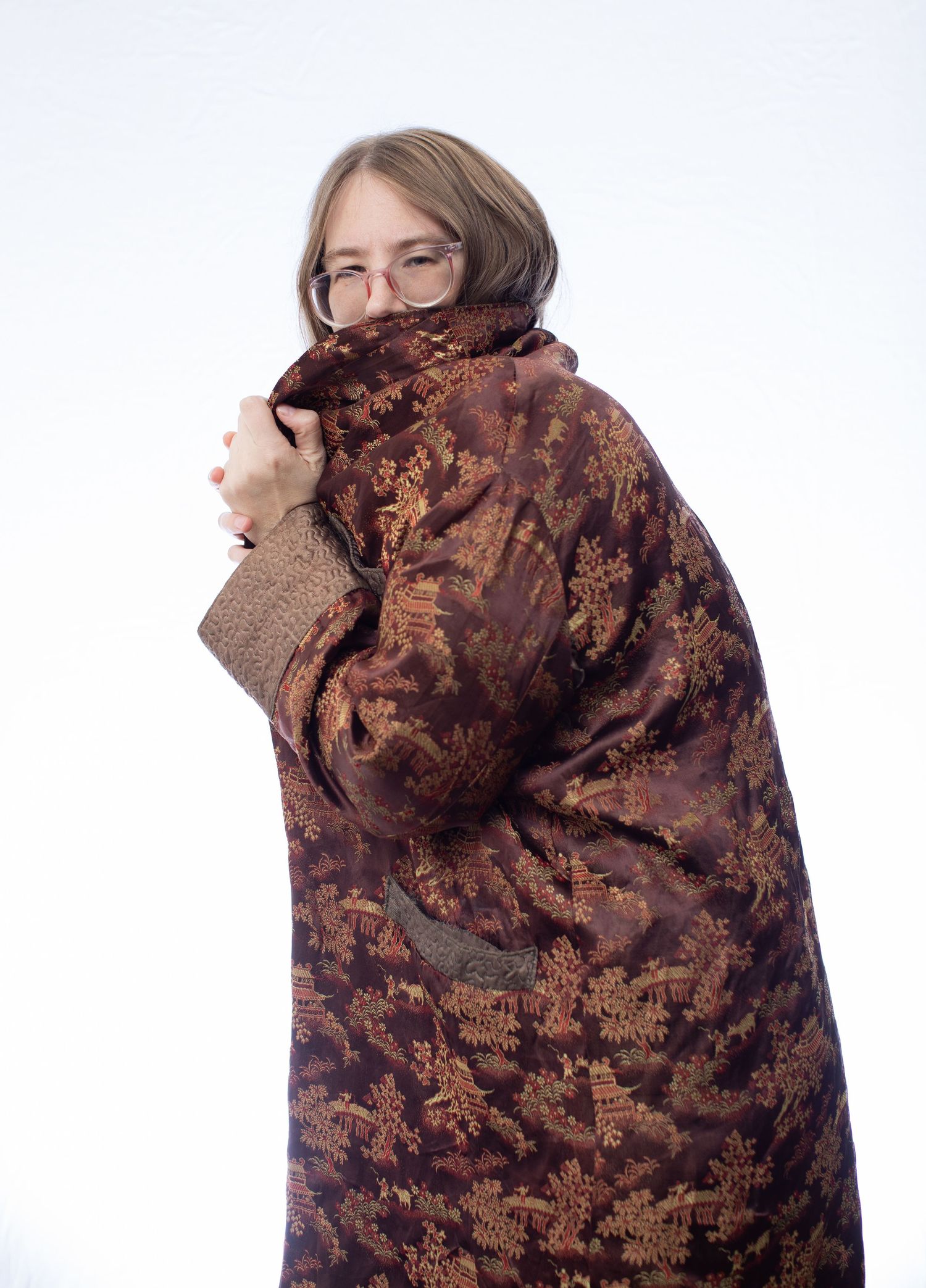 Person wrapped in brown patterned blanket peering over shoulder while wearing glasses.