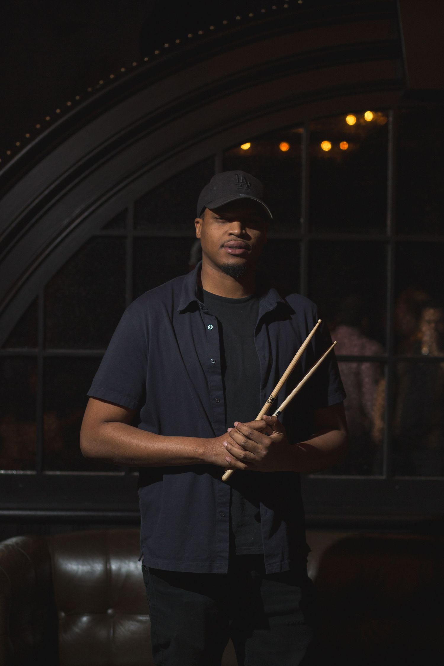 A drummer holds drumsticks while posing in front of an arched window with ambient lighting.