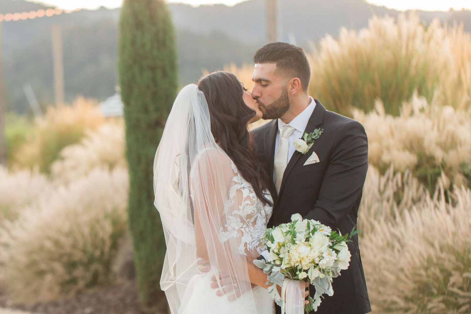 Newlyweds share a romantic moment outdoors with a white floral bouquet against golden light.