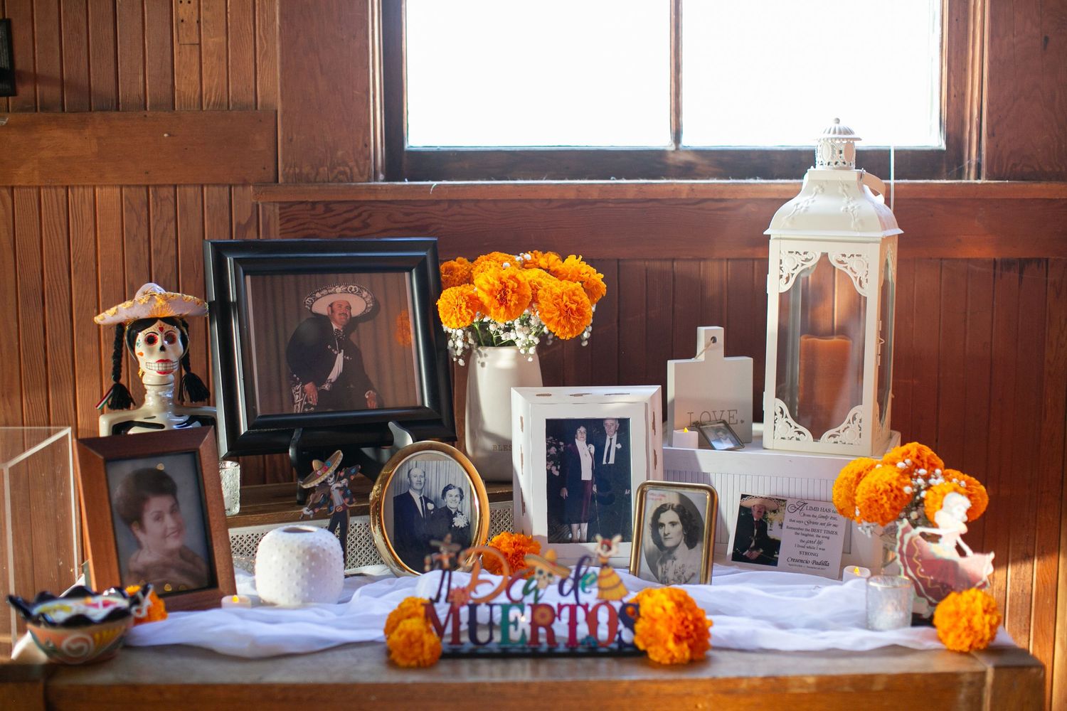 Memorial table display with framed photos, orange flowers, and lanterns in rustic wooden setting.