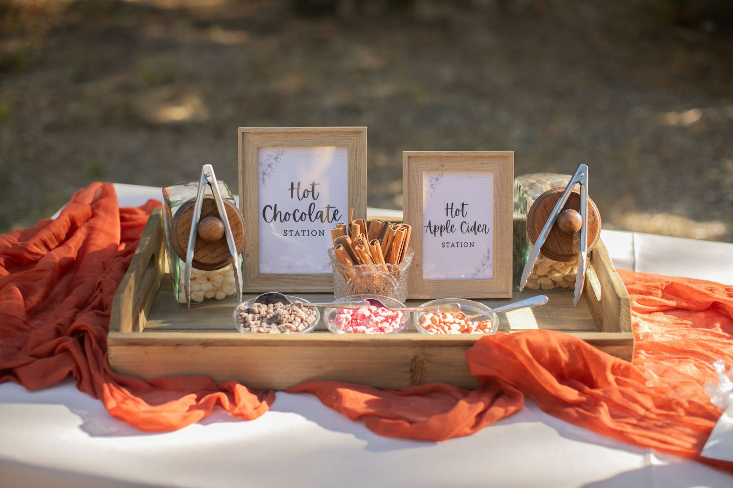 Hot chocolate and cider station with rustic signage and orange fabric decor.