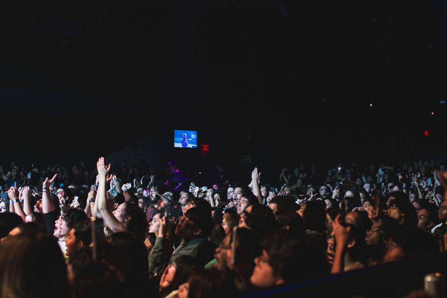 Concert crowd raises hands in darkness with distant stage lights and video screen visible.