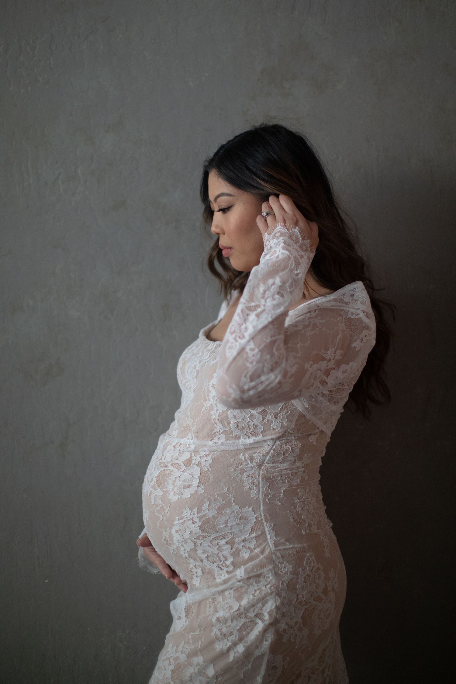 Side profile of pregnant woman in white lace dress against gray wall.