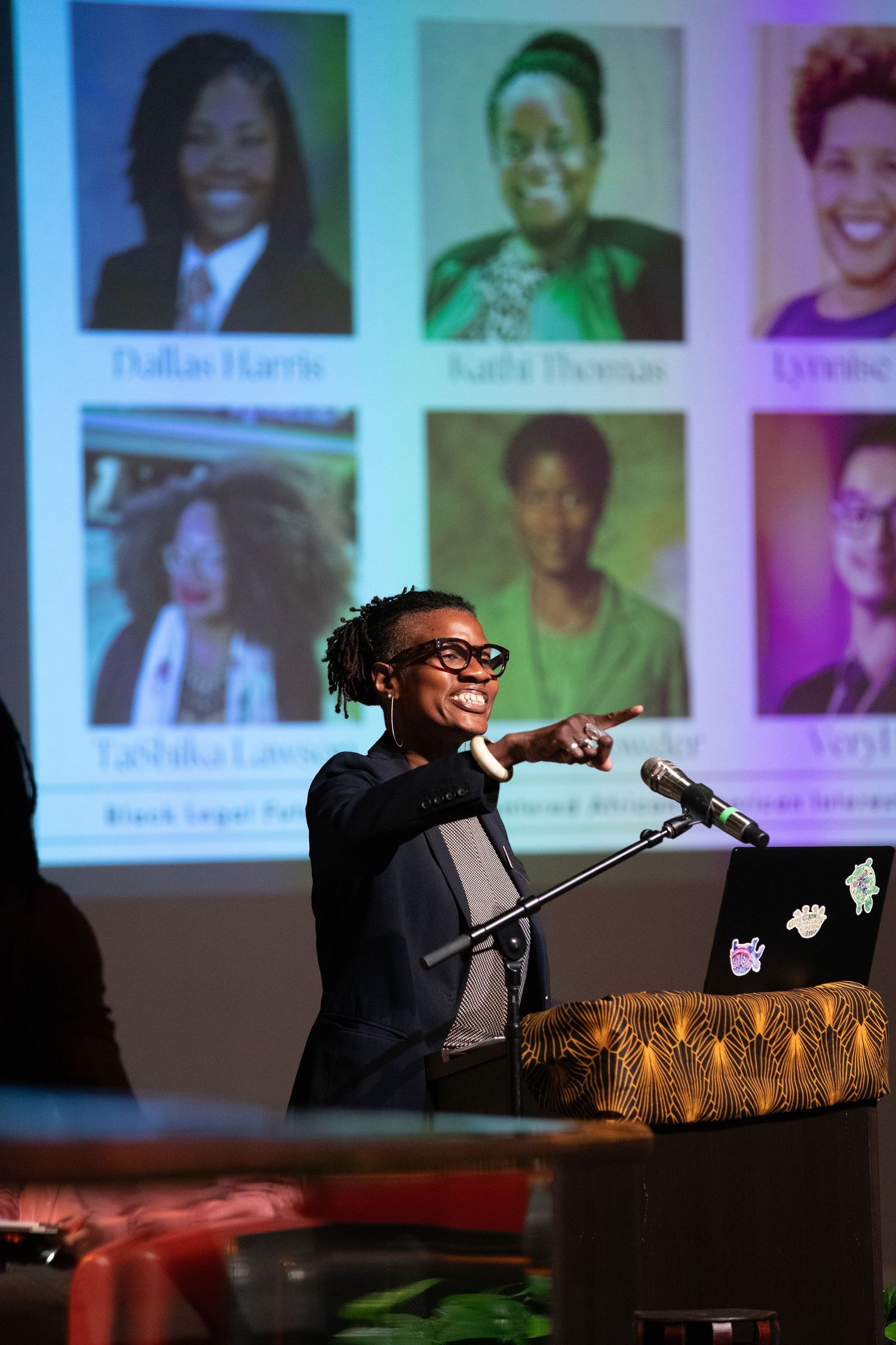 Speaker at a podium gestures animatedly during presentation with biographical photos projected on screen behind them.