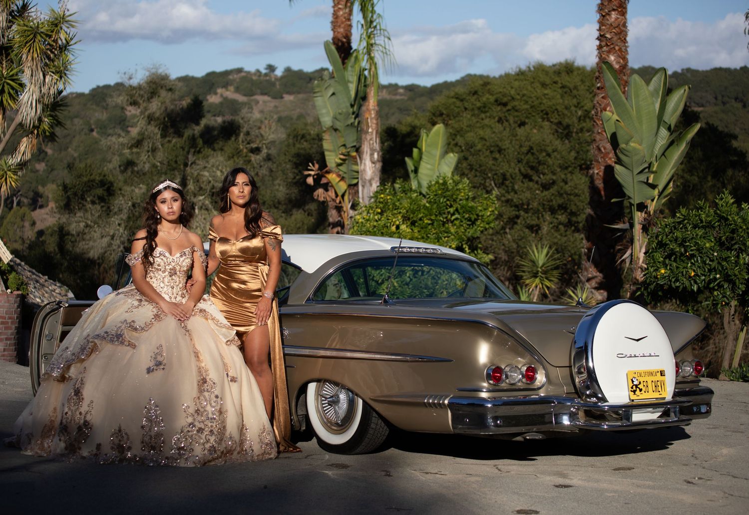 Two people in formal attire pose beside a vintage bronze car in a sunny outdoor setting.