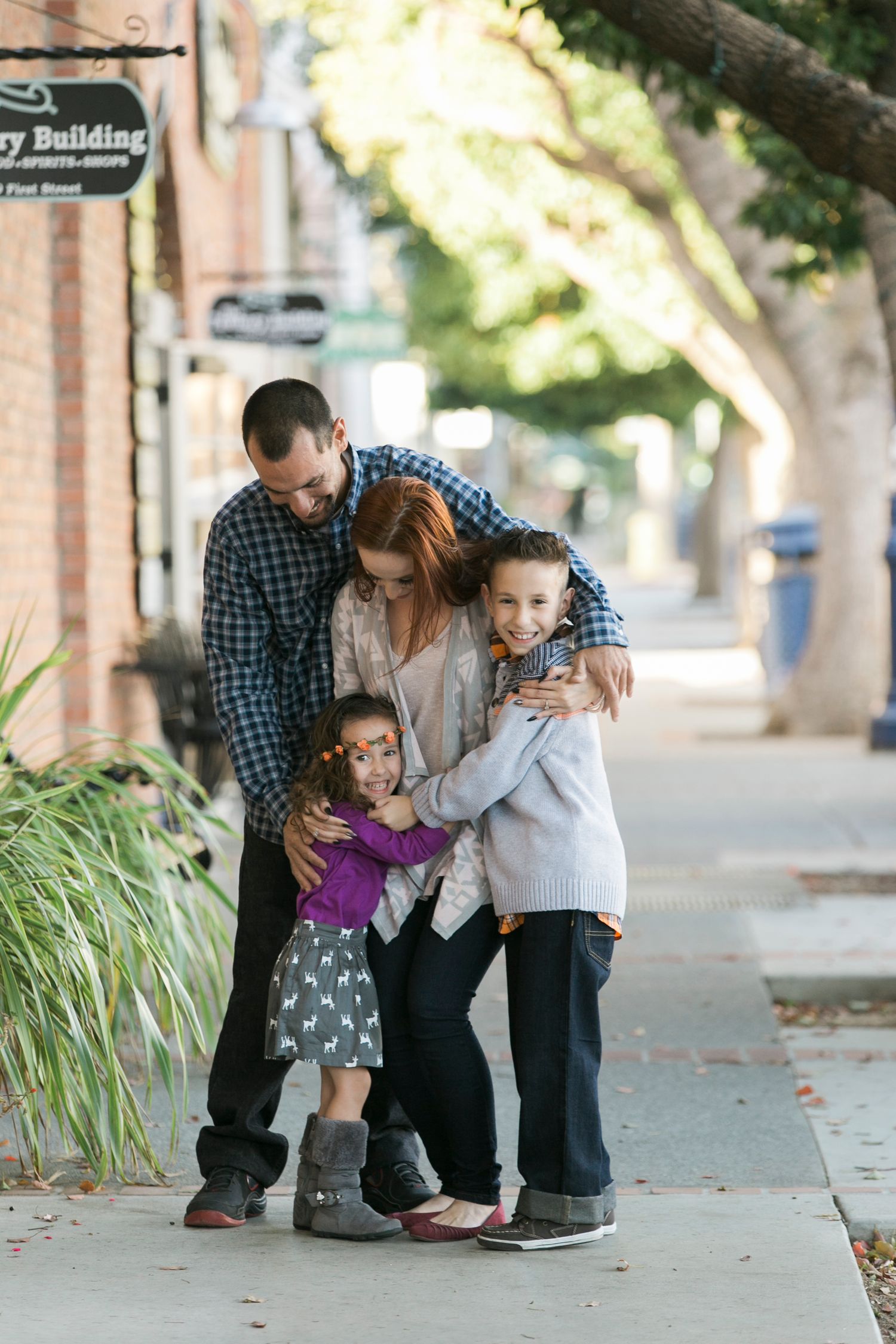Sweet family moments captured on a city sidewalk with brick buildings and landscaping.