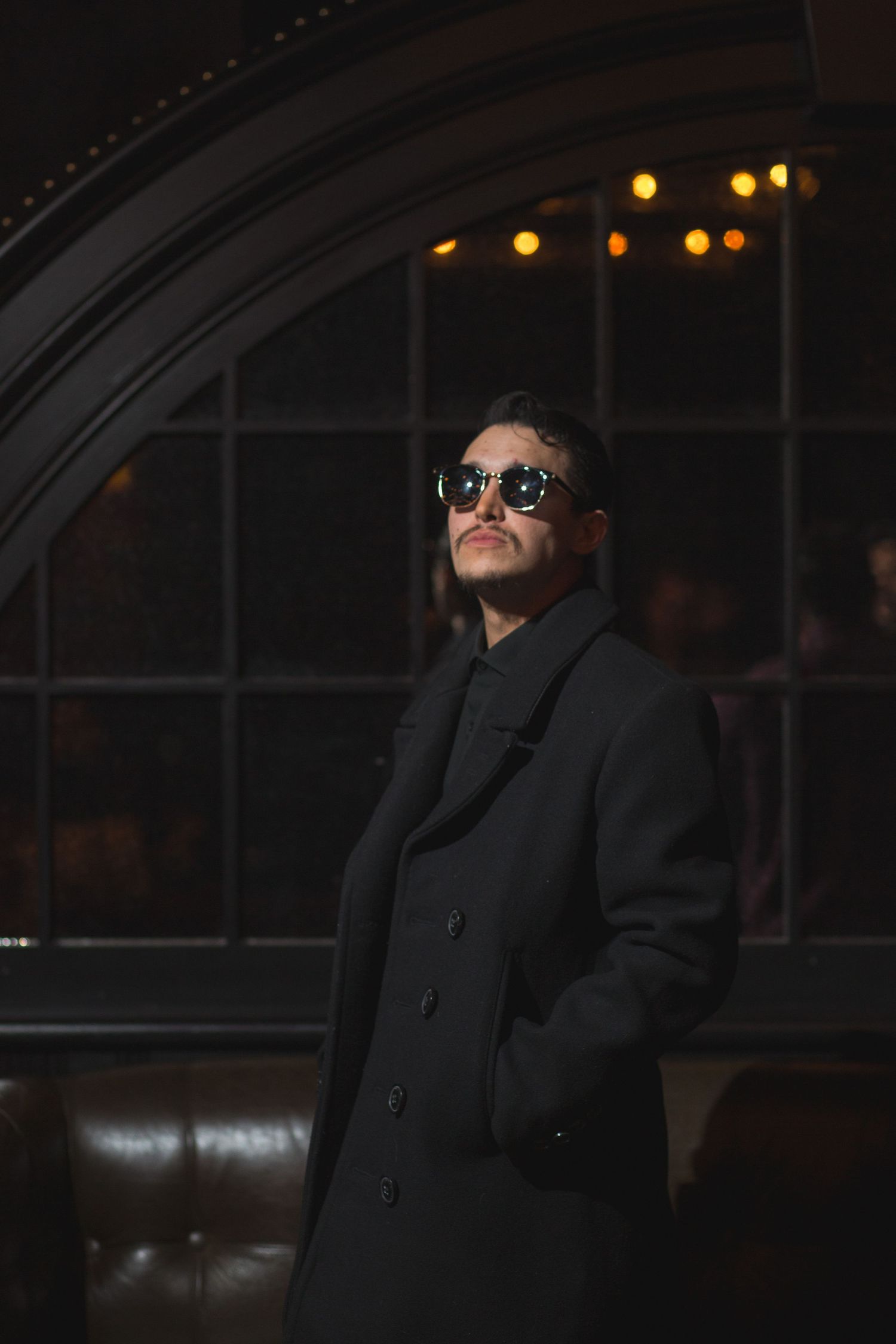 A stylishly dressed performer in sunglasses and long coat poses in a dimly lit venue.