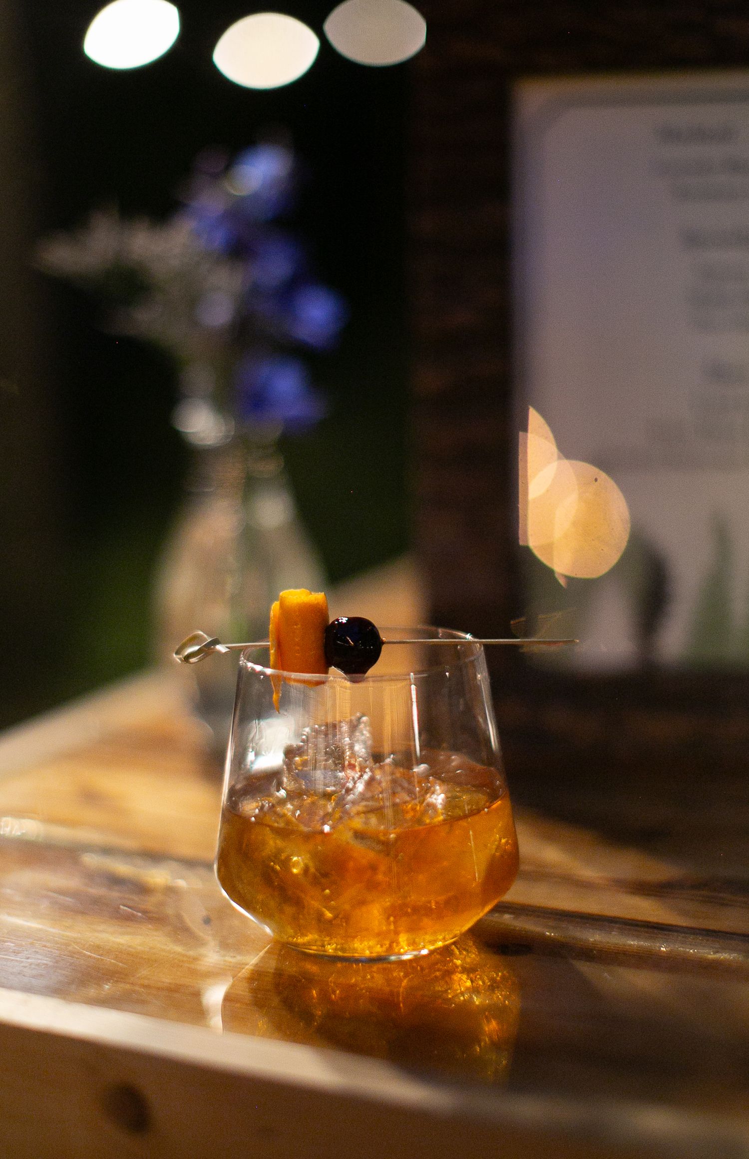 Amber-colored cocktail with garnish in a stemmed glass against a dark background with blue flowers.