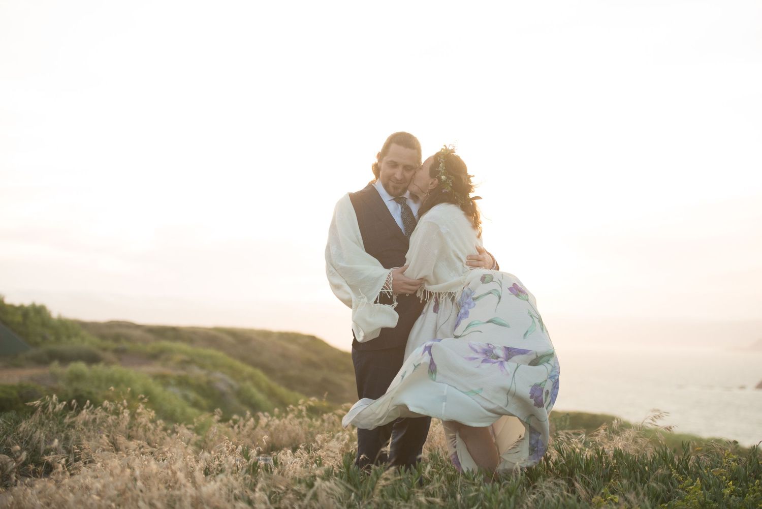 At sunset, a couple in flowing white clothes embraces along California's coastal cliffs by the ocean.