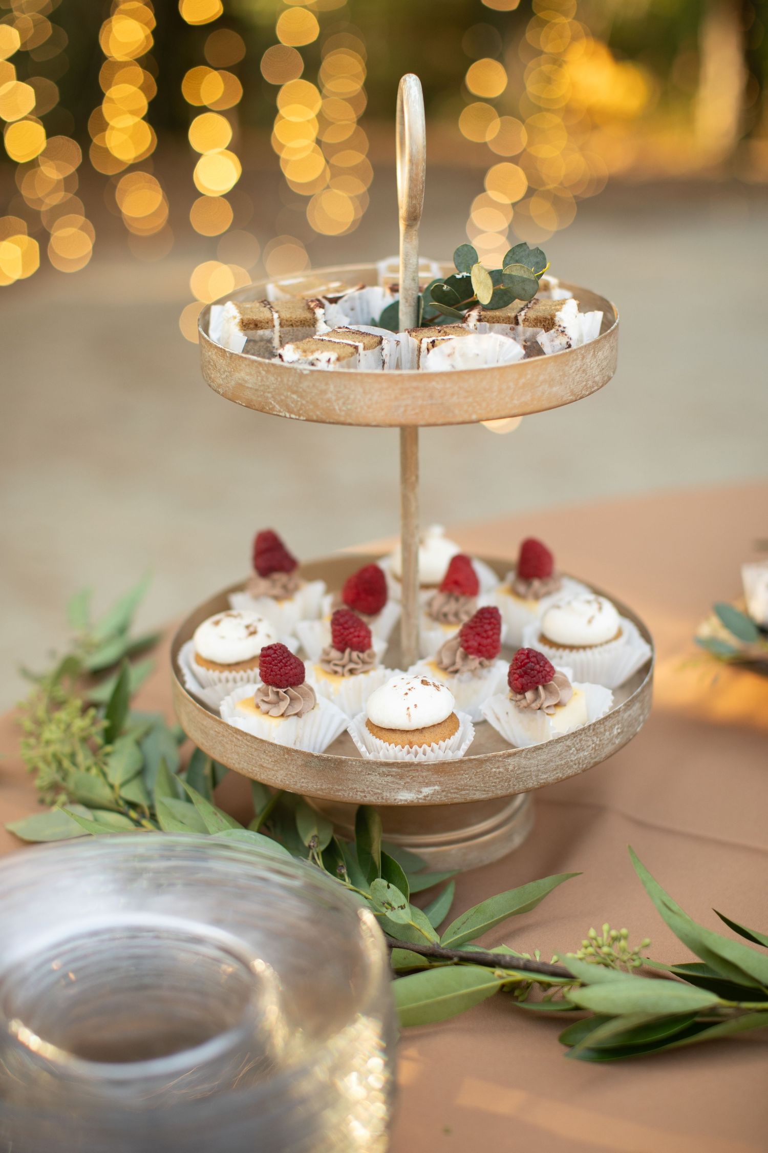 Elegant dessert display with cupcakes on tiered stands decorated with greenery and warm bokeh lighting.