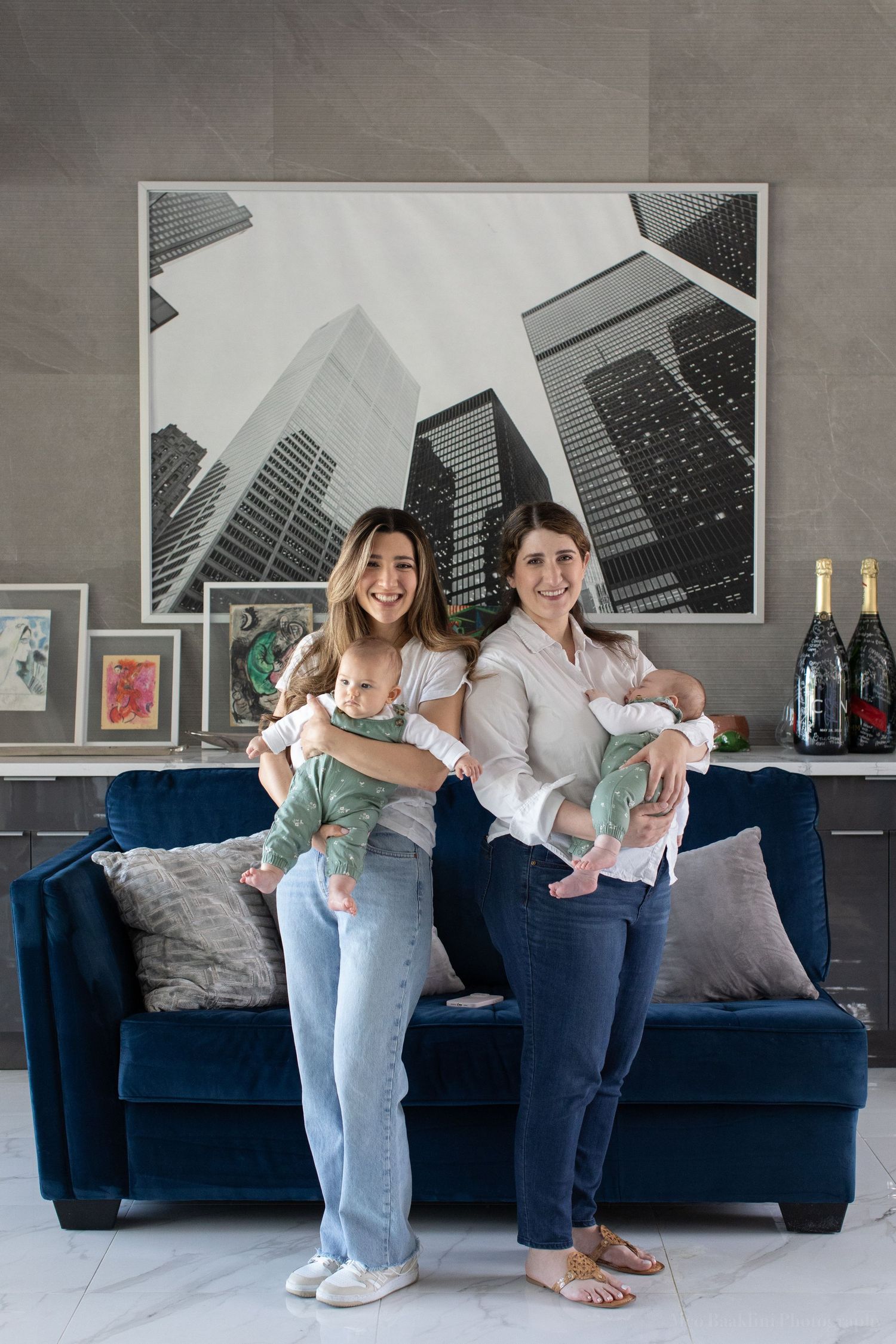 Two people pose with babies beside a navy couch in a modern apartment with city skyline artwork.