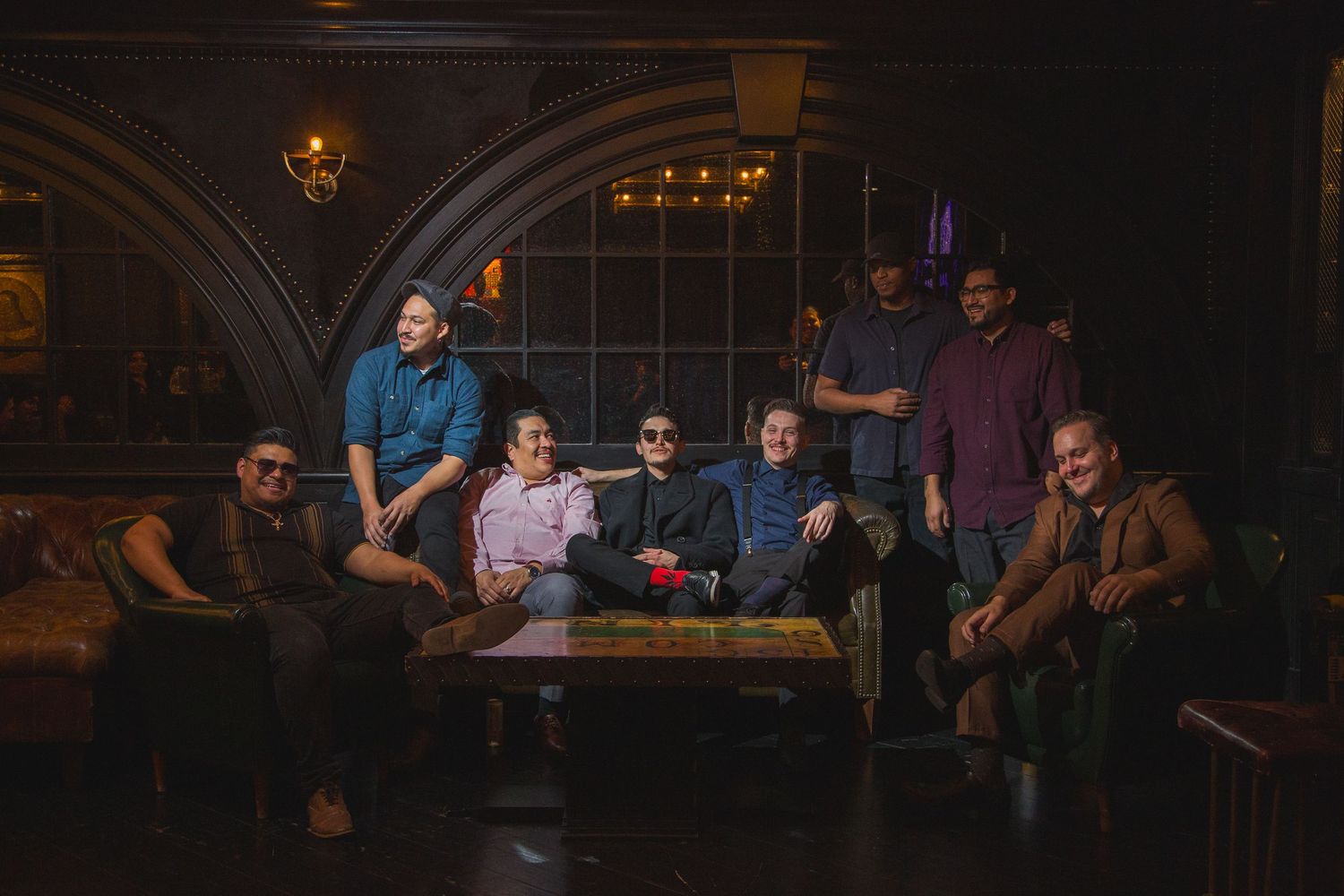 A group of musicians pose together on leather seating in a darkened venue with arched architecture.