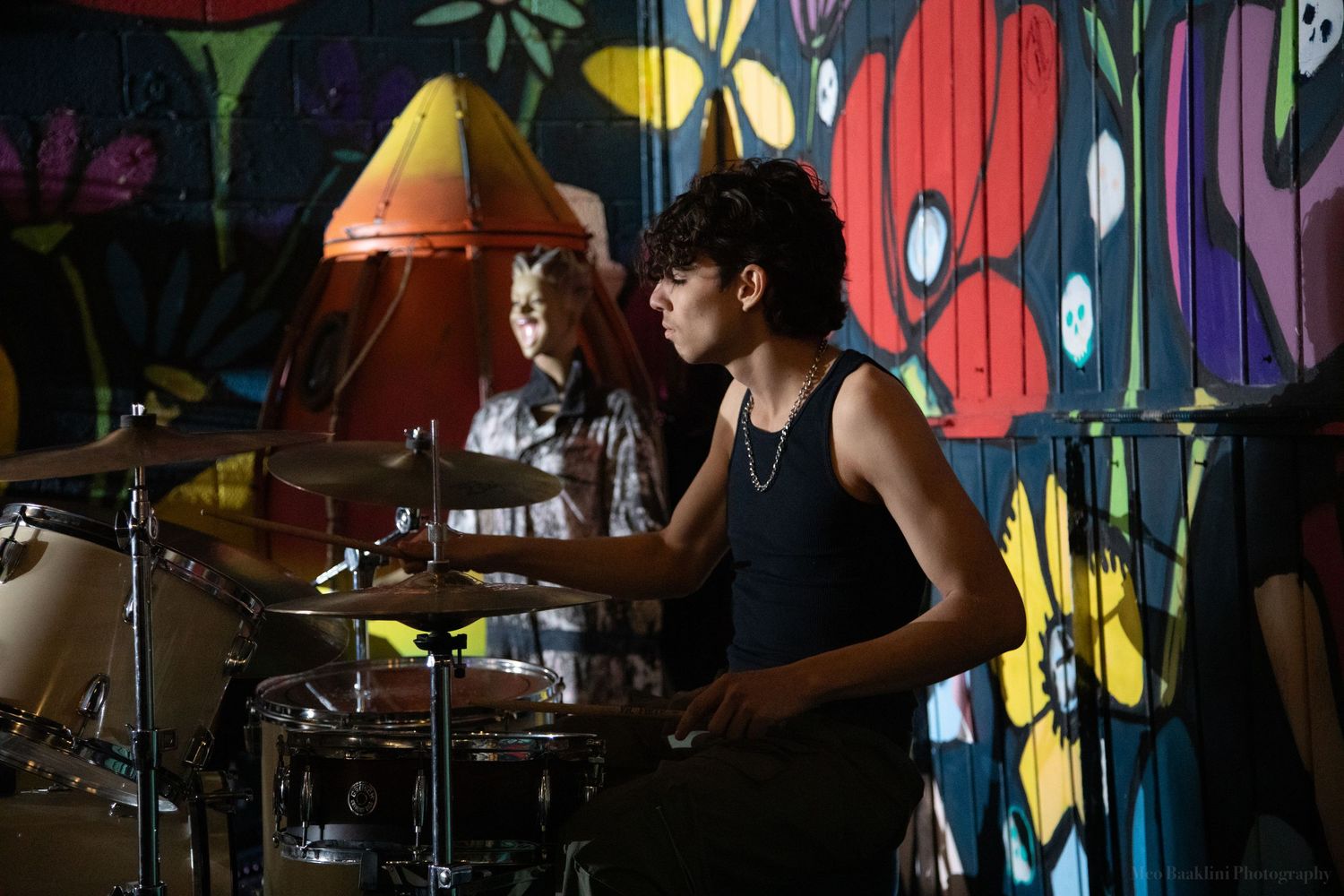 Musicians perform in front of a colorful graffiti wall with drums and musical equipment visible.