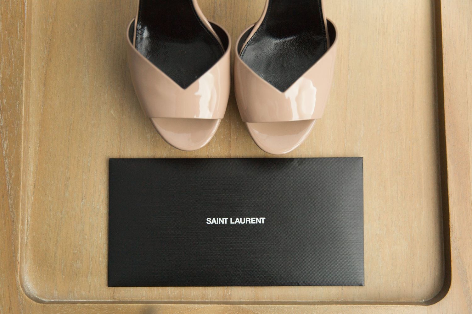 A pair of pale pink patent leather peep-toe shoes displayed above a black Saint Laurent box on beige surface.