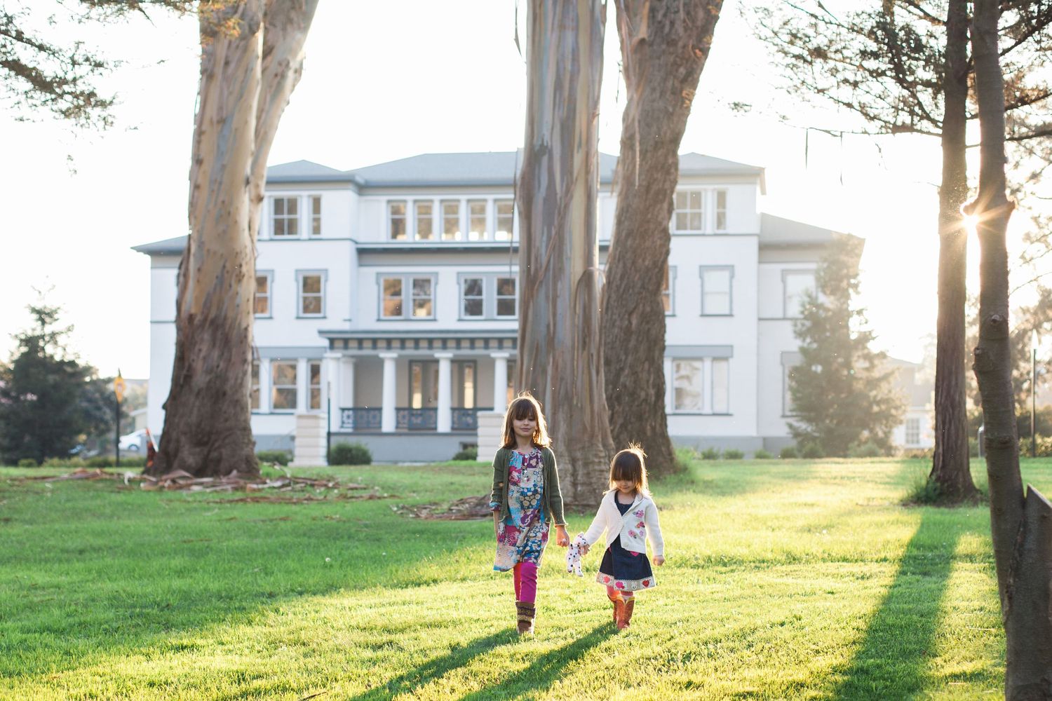 Two children walking on grass path in front of large white Victorian mansion at sunset.