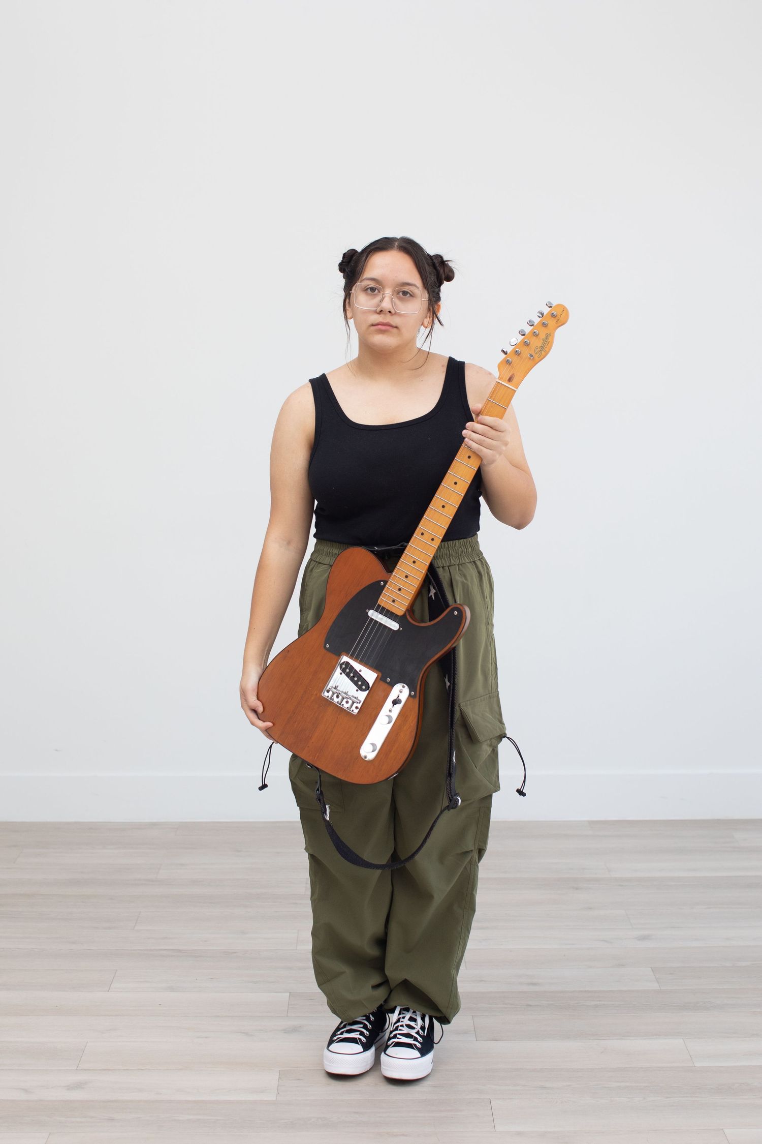 Musician holds brown electric guitar while wearing casual attire against white backdrop.