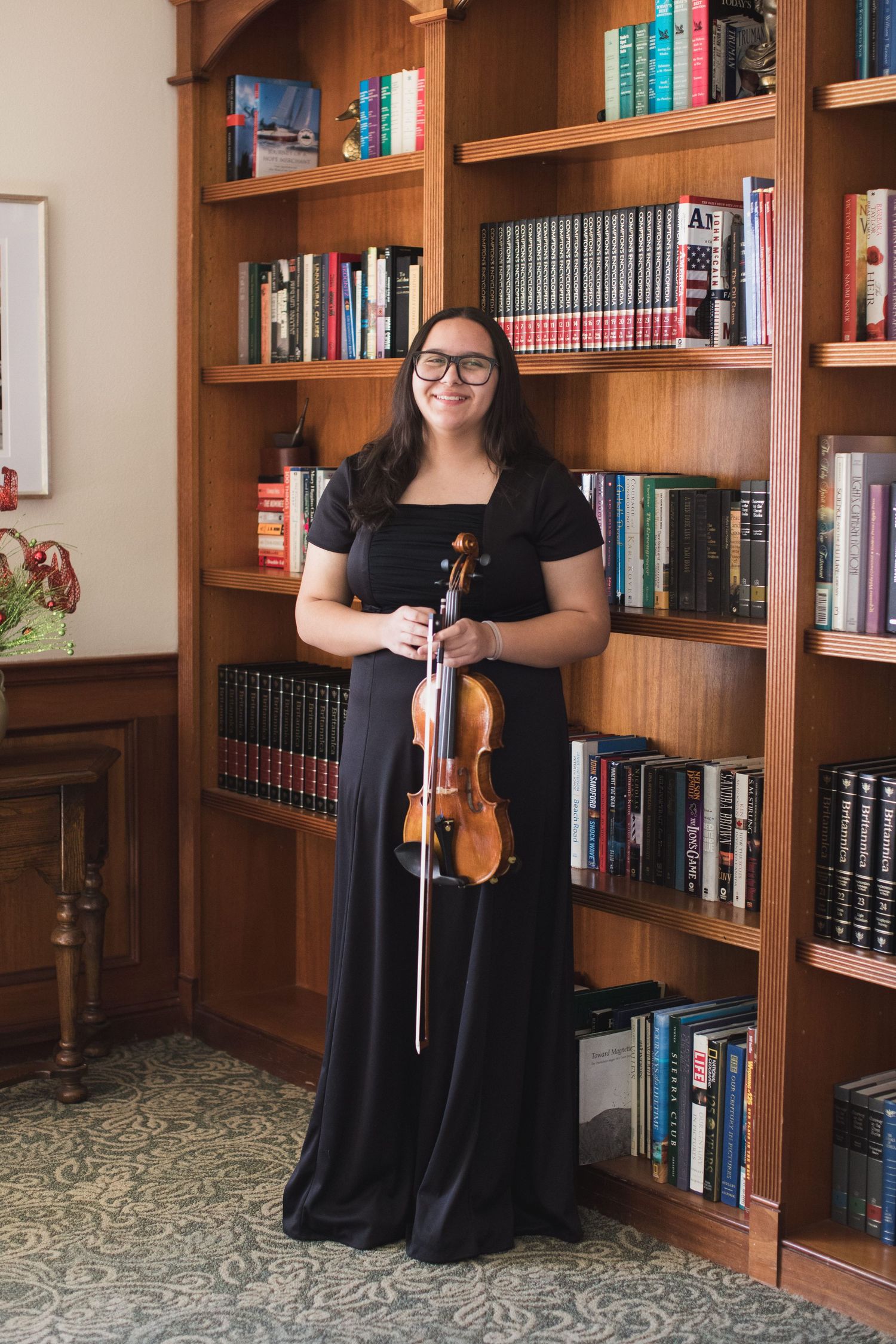 Musician poses with violin in front of wooden bookshelf filled with books.