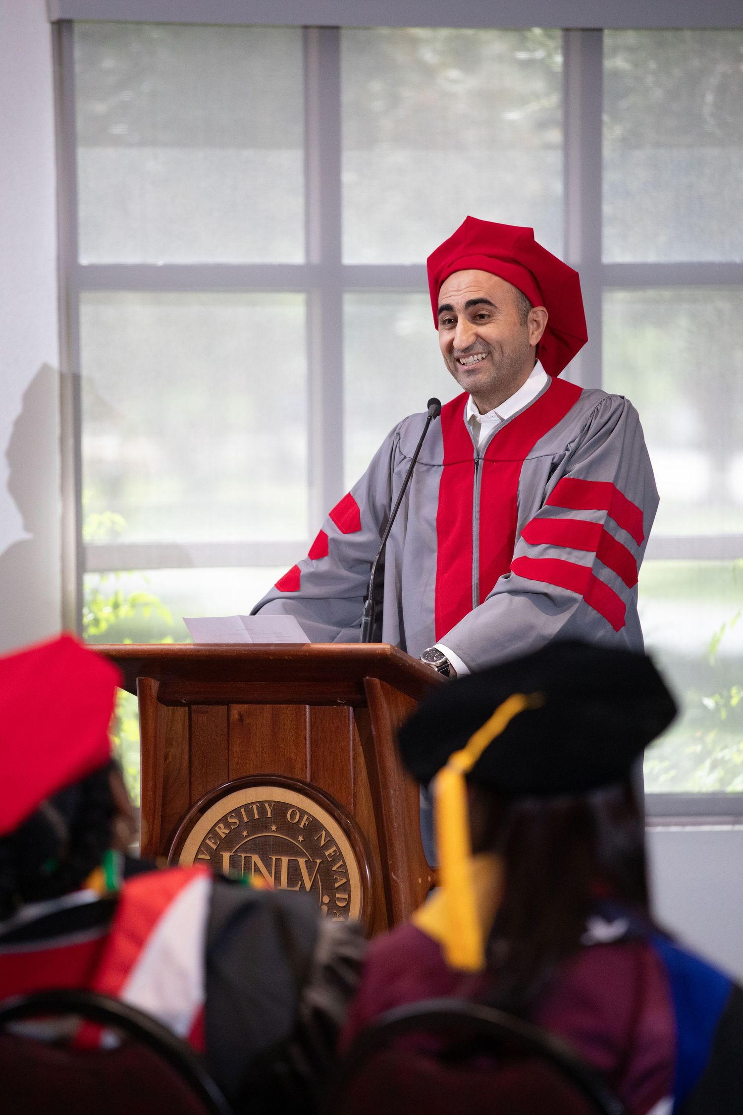 Speaker in red and grey doctoral regalia addresses audience from wooden UNLV podium.