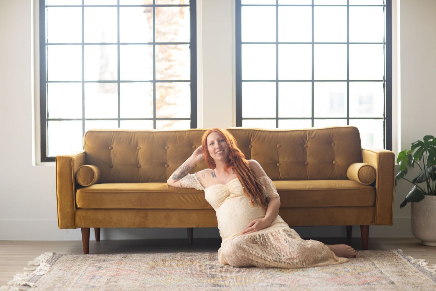 Pregnant woman in cream dress relaxing on brown leather couch near windows.