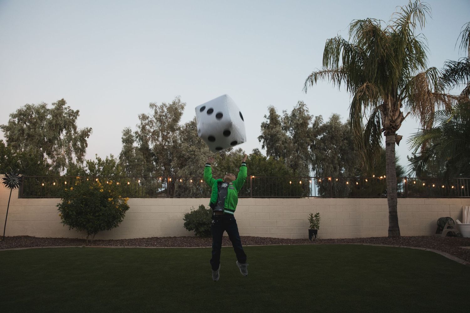 Person in green shirt playing with large inflatable dice in a backyard with string lights.