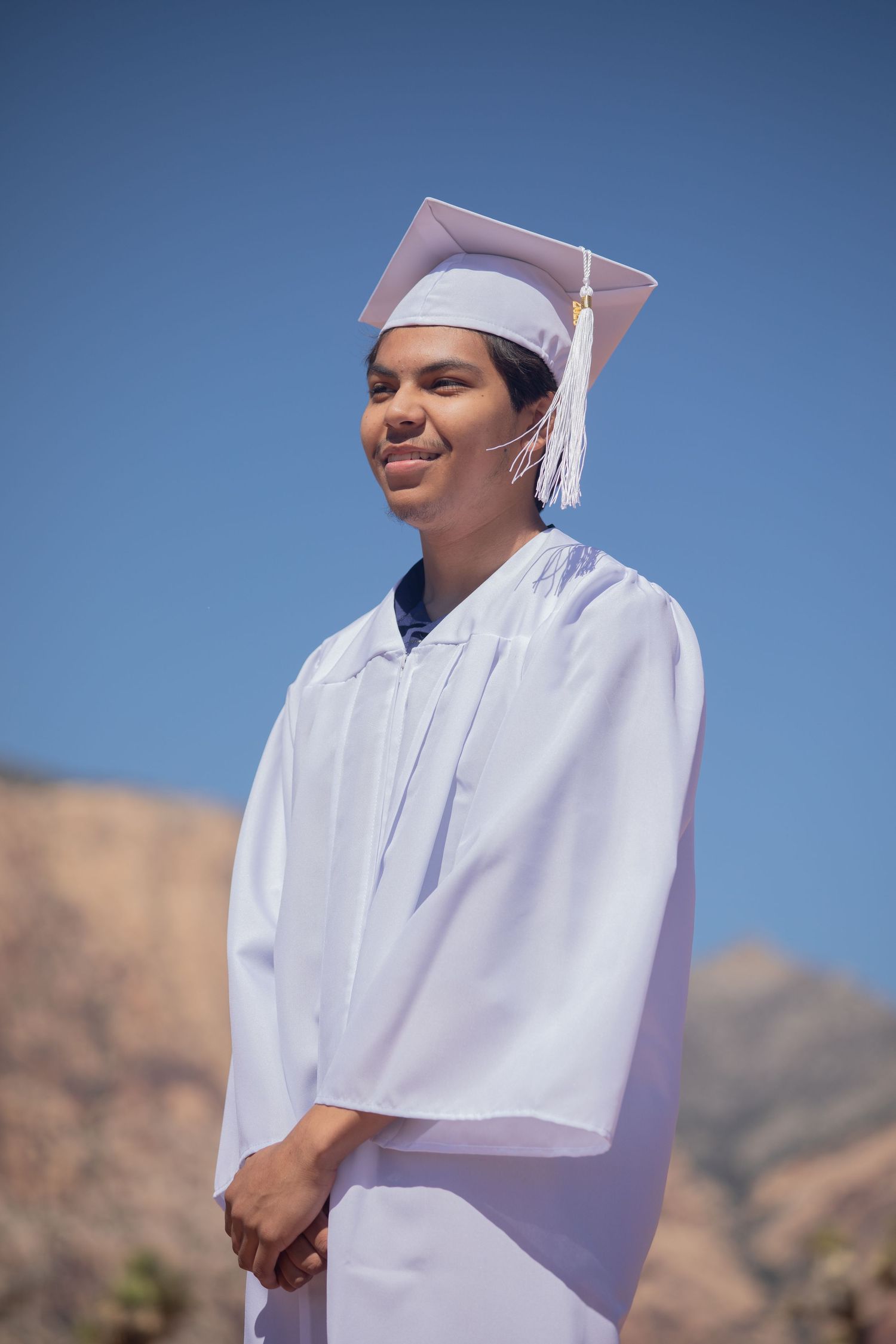 Graduate in white cap and gown poses against mountain landscape on sunny day.