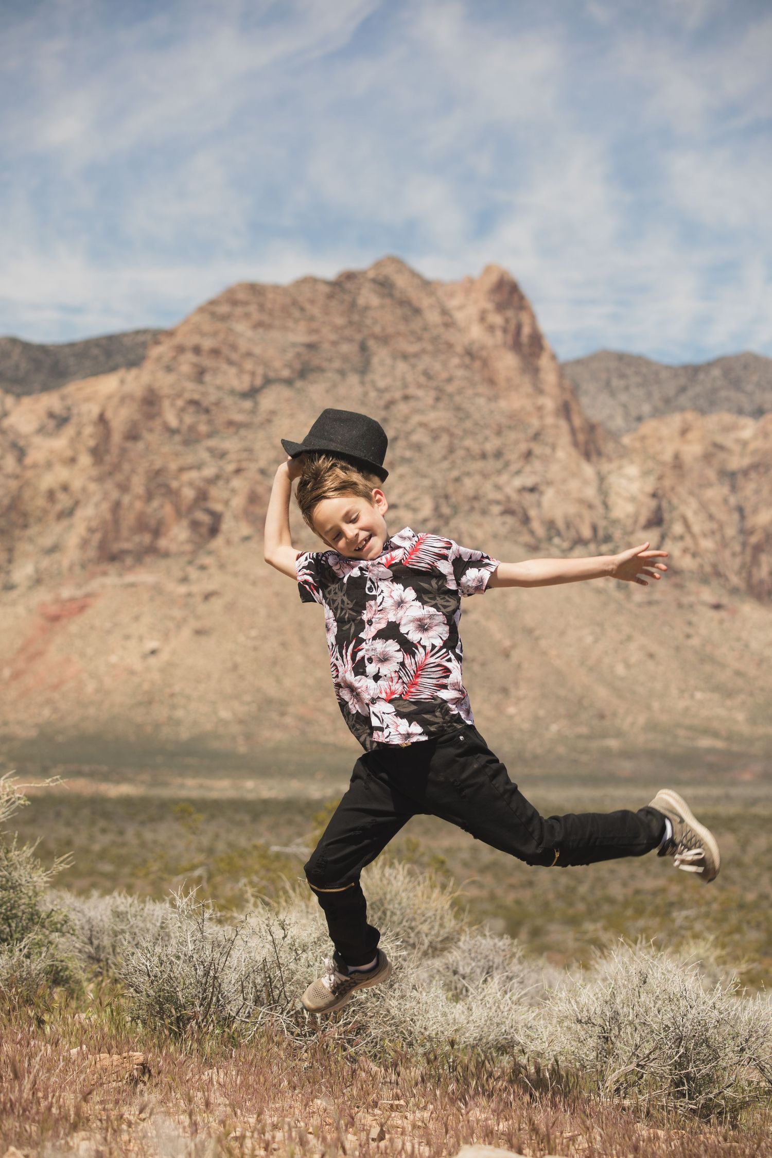 Dancing figure in black pants and patterned shirt leaps joyfully against desert mountain backdrop.