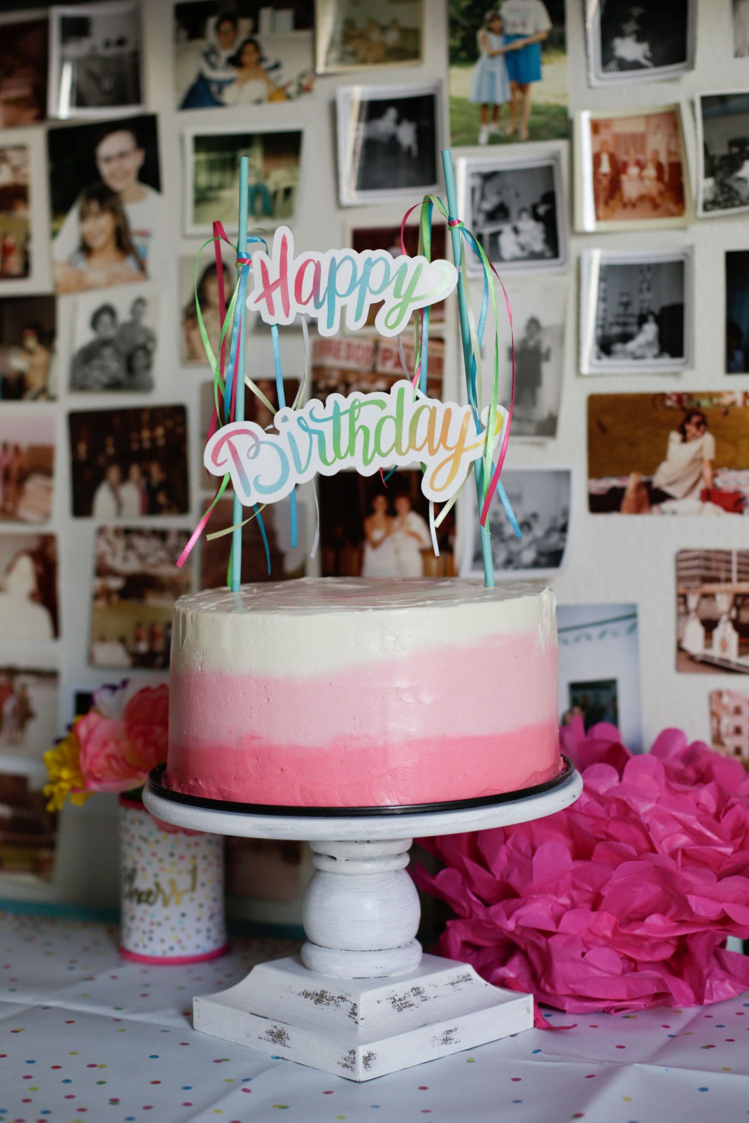 White birthday cake with pink ombre frosting displayed on stand with vintage photos in background.