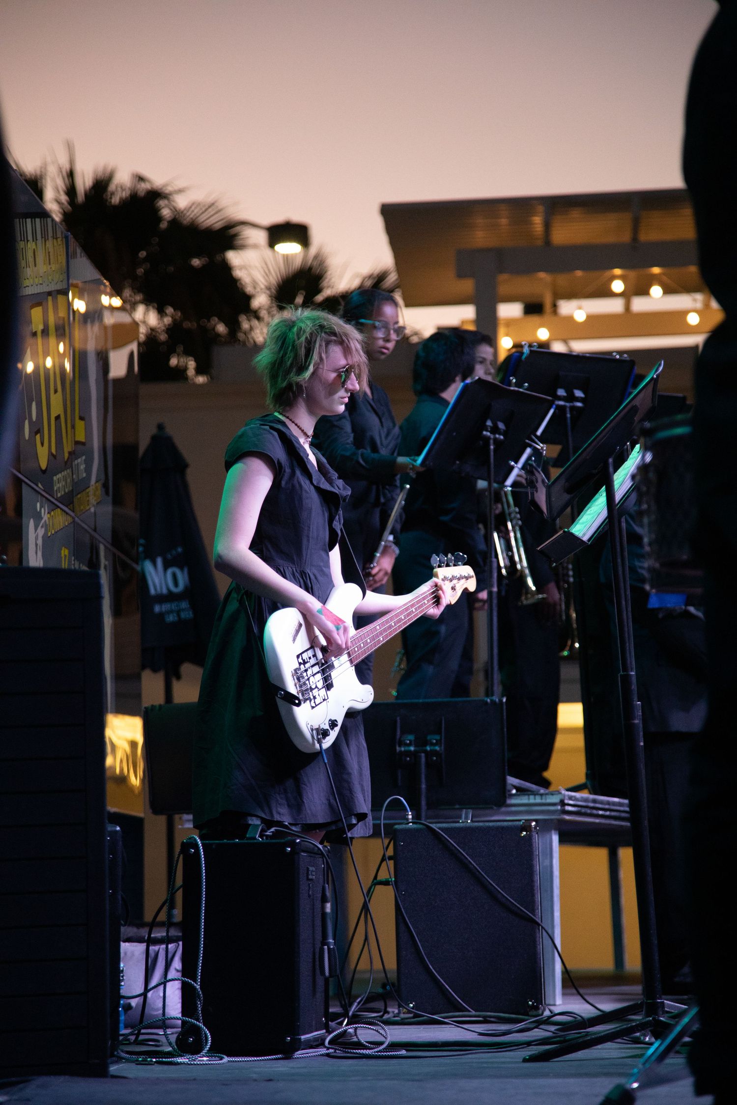 Musician with colorful hair plays bass guitar on outdoor stage at twilight.