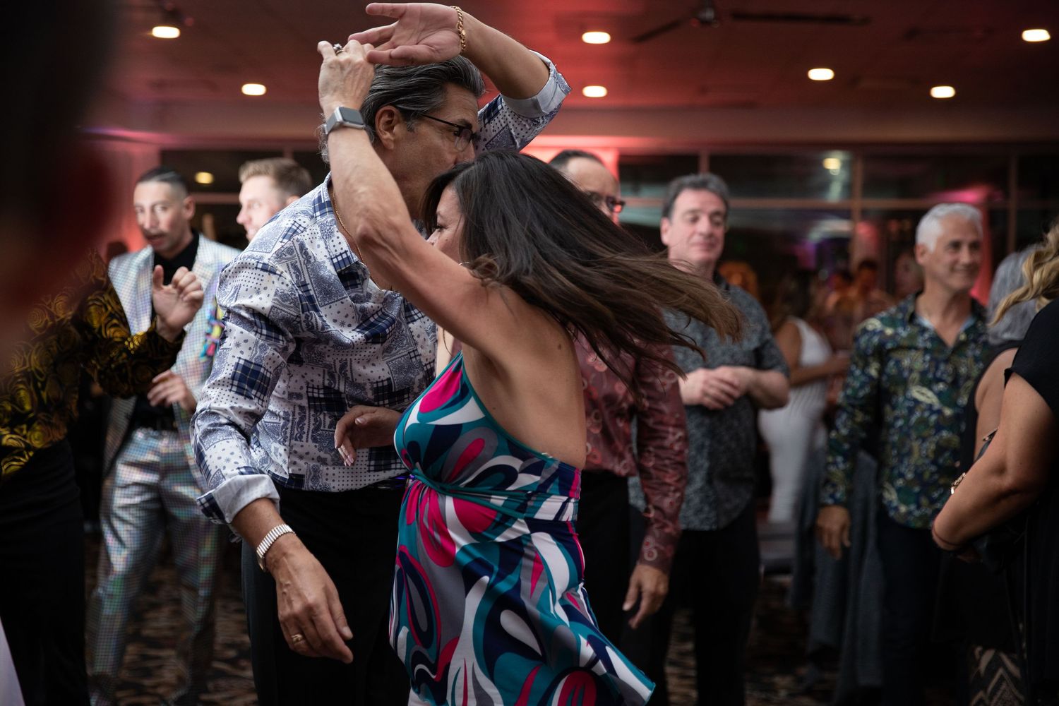 Dancers move energetically on a dimly lit reception dance floor with red lighting.
