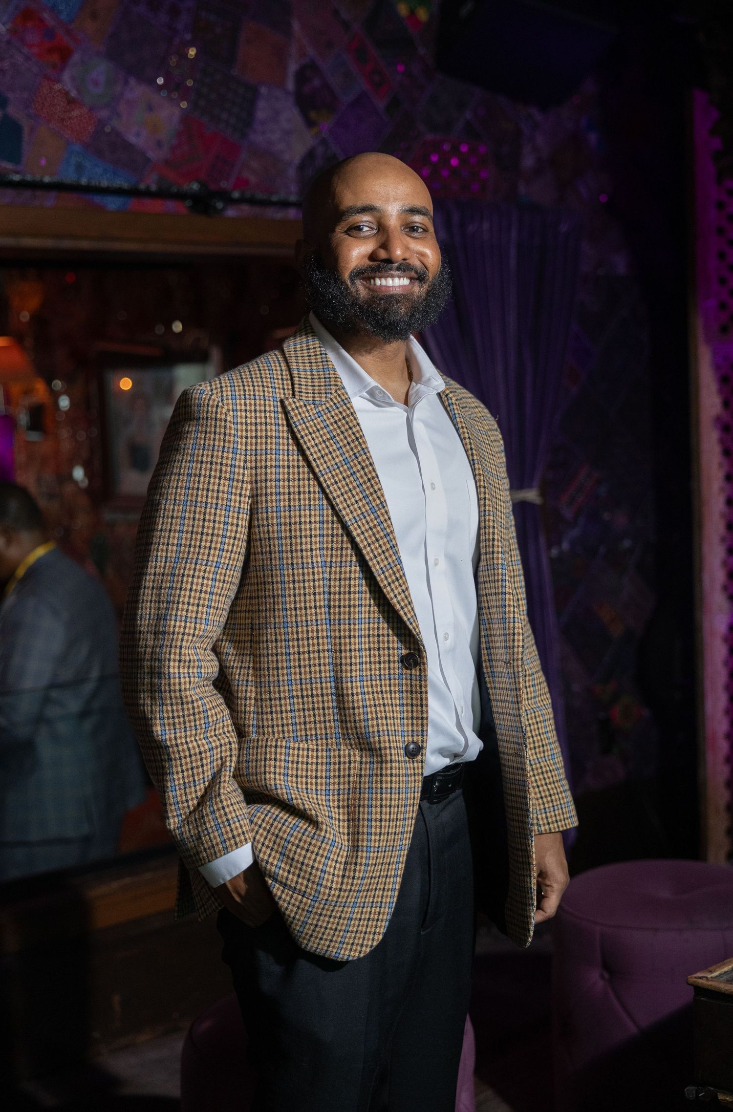Professional wearing a plaid blazer and light colored shirt stands smiling in a dimly lit venue.