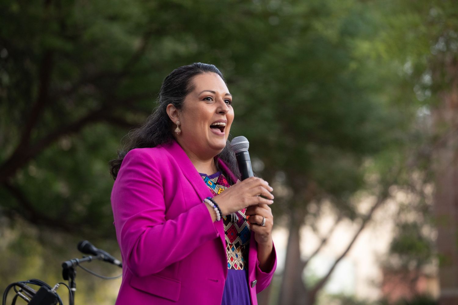 A speaker in a bright pink blazer addresses an outdoor audience with enthusiasm using a microphone.
