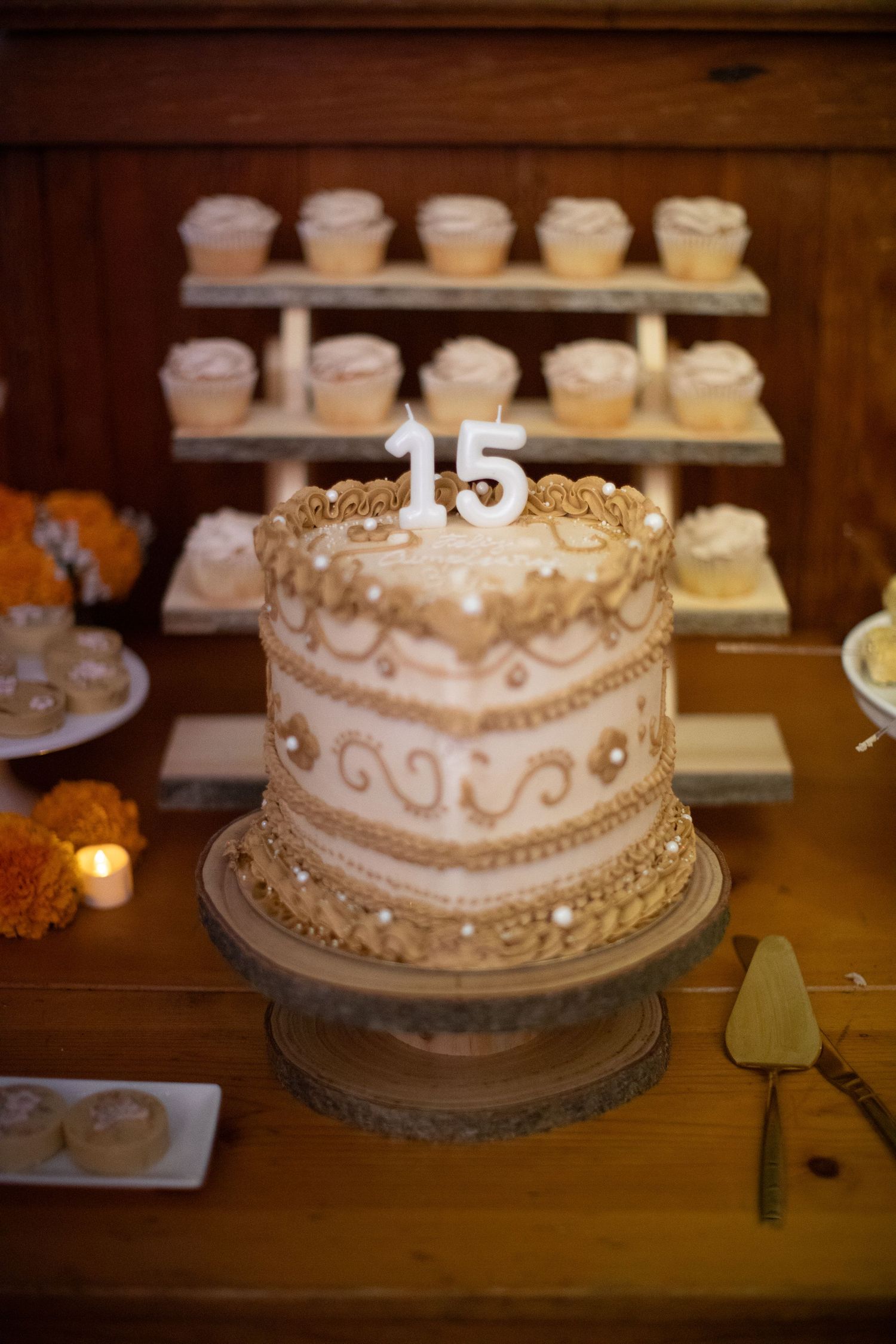 An elegant tiered white and gold cake decorated with the number 15 and surrounded by cupcakes.