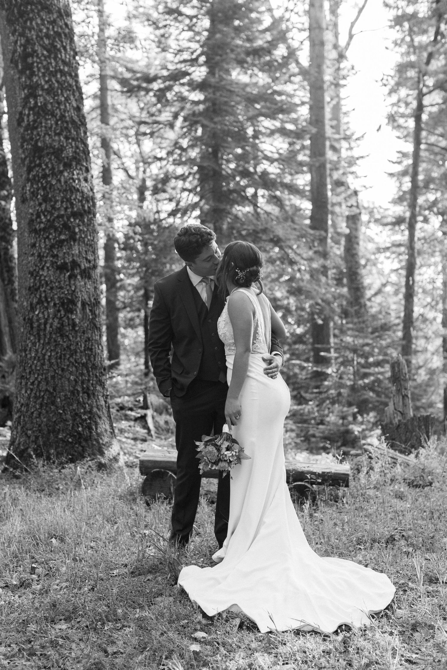 A romantic black and white wedding photo of a couple in formal attire standing among tall forest trees.