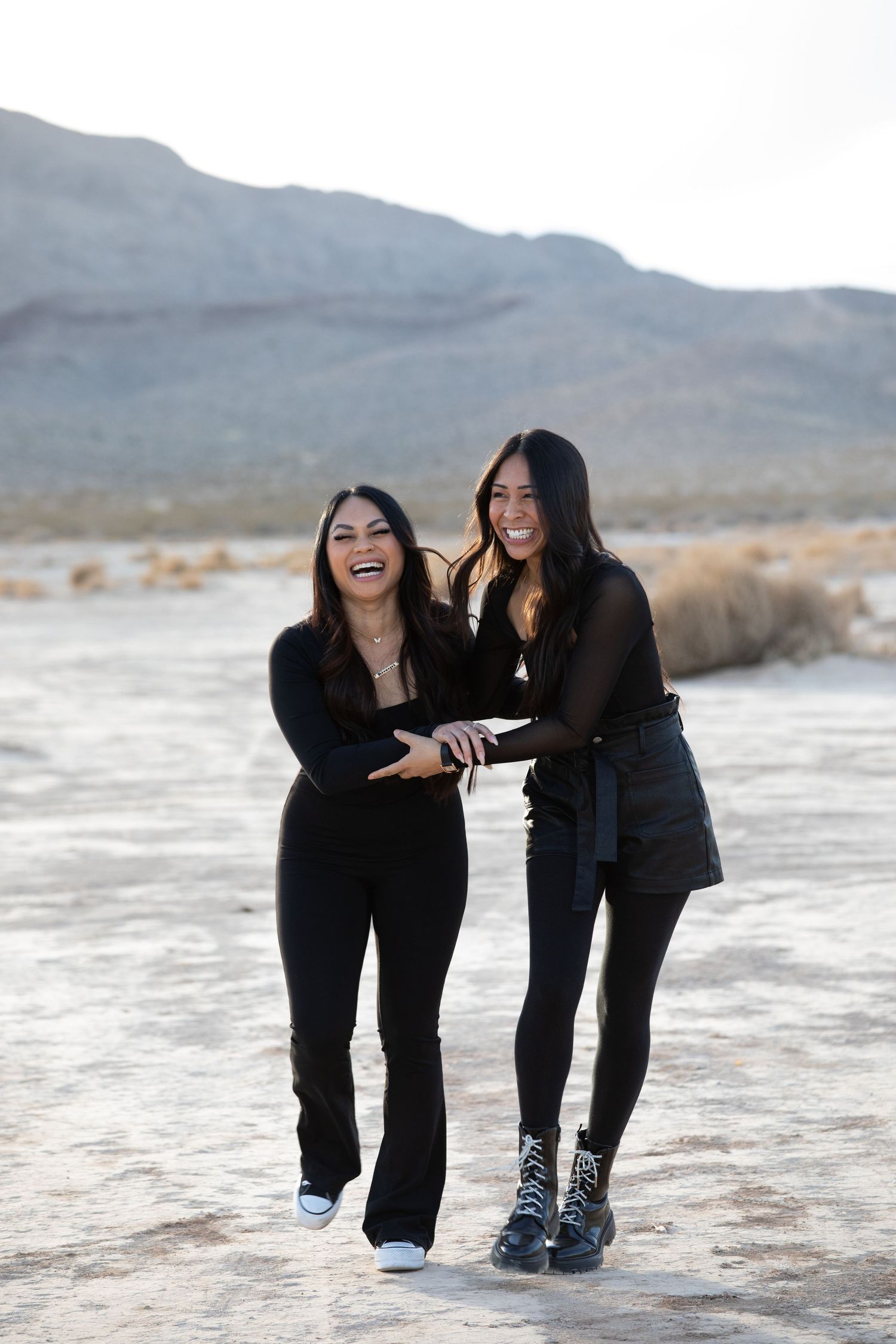 Two people in black outfits share a candid laugh together in a desert landscape.