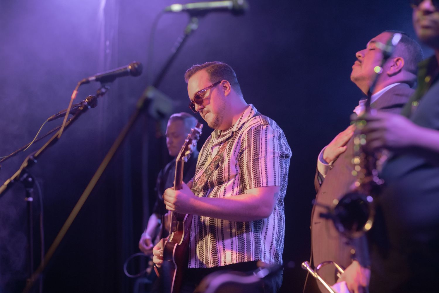 Musicians perform on stage with guitar and instruments under purple stage lighting.