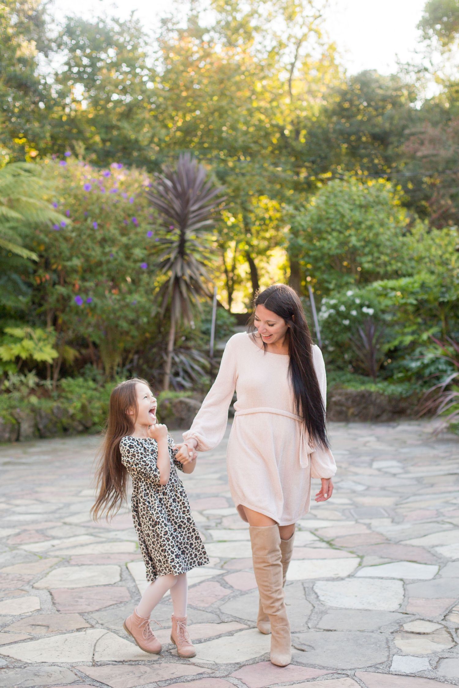 Mother and daughter in matching neutral tones dance on stone pathway in garden setting.