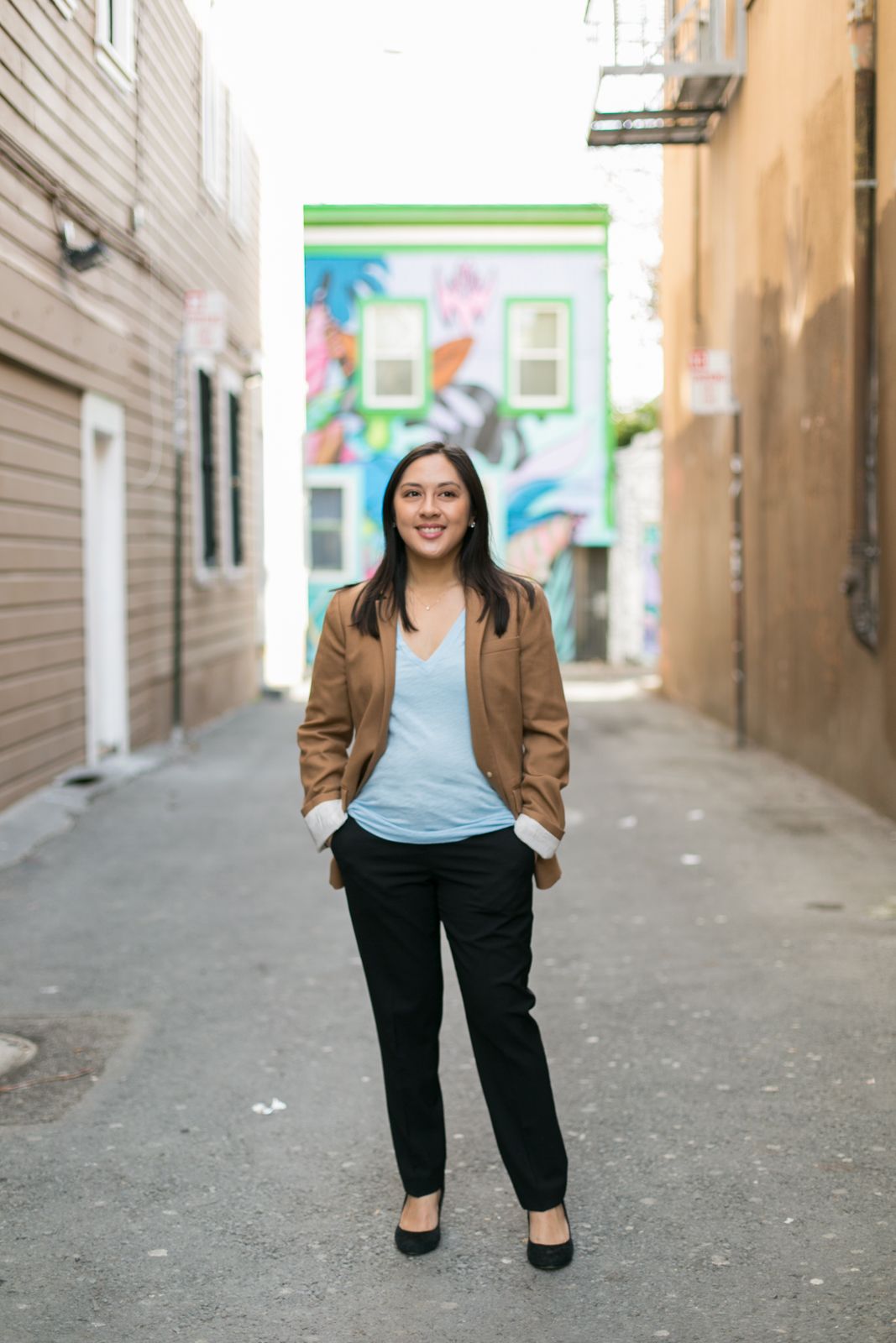 Professional portrait in tan blazer and casual attire standing in urban alley with colorful backdrop.