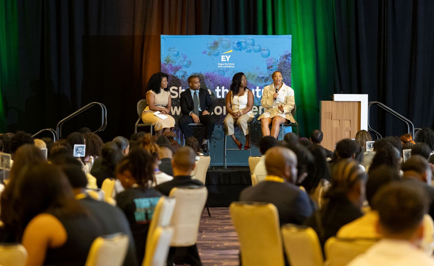 Large audience seated in a dimly lit conference room with green stage lighting.