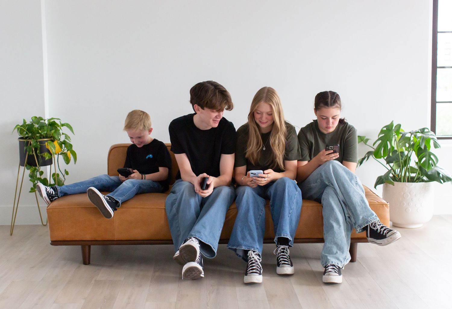 Four people relaxing on a brown couch with house plants in a minimalist white room.