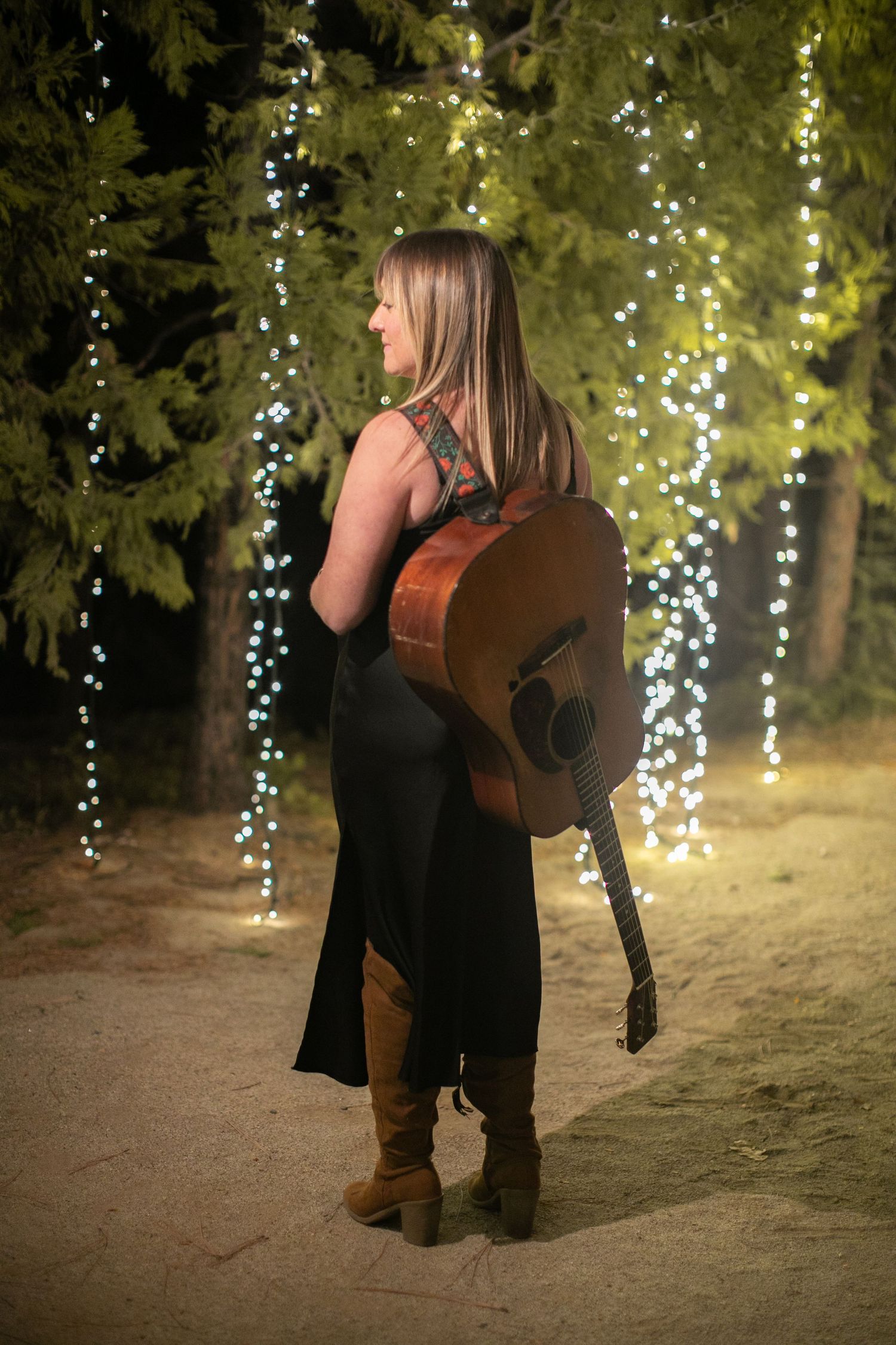 Musician with acoustic guitar photographed against twinkling fairy lights in outdoor setting.