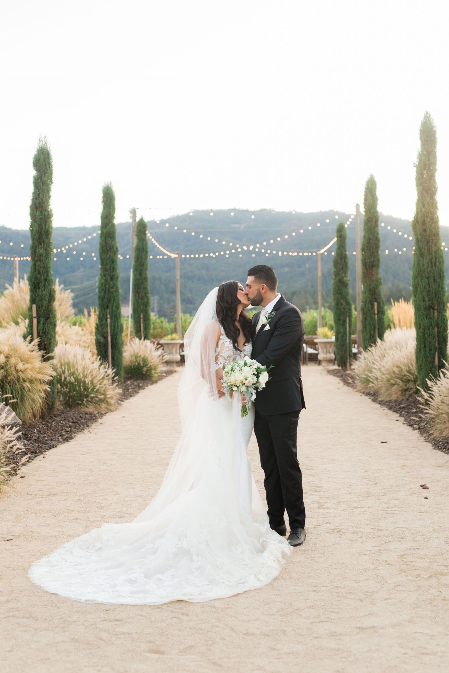 Couple embraces on a garden path lined with cypress trees and string lights at dusk.