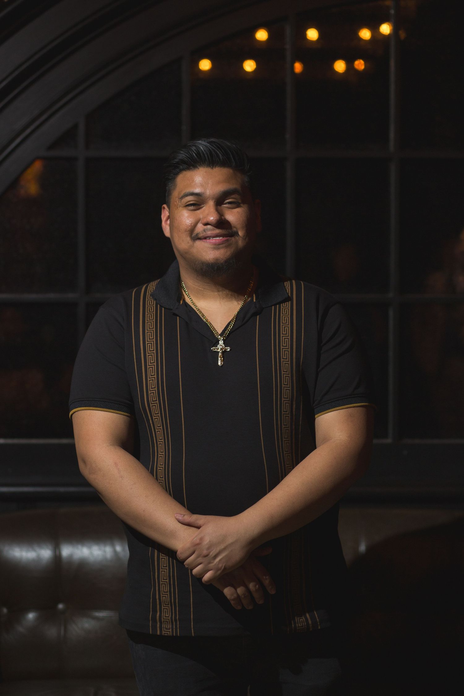 A performer in a striped shirt smiles warmly in front of an arched window with ambient lighting.