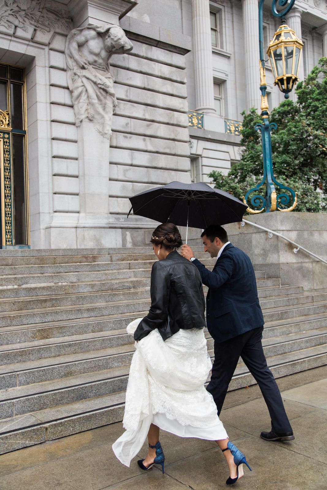 A couple walks up stone steps in formal attire sharing a black umbrella on a rainy day outside an elegant building.