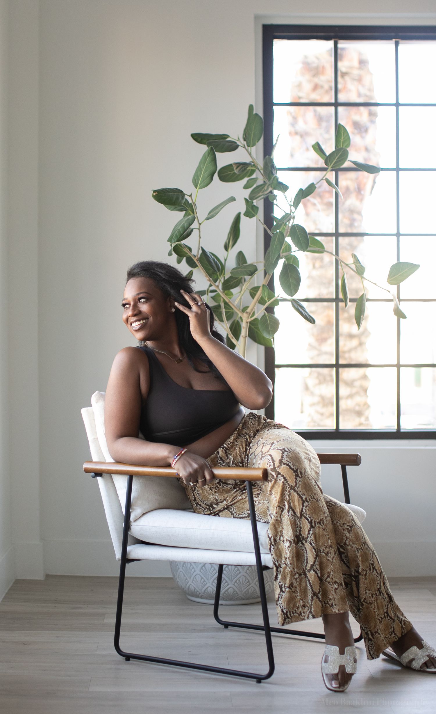 Stylish person in black top and snake print skirt poses in modern chair by window with plant.