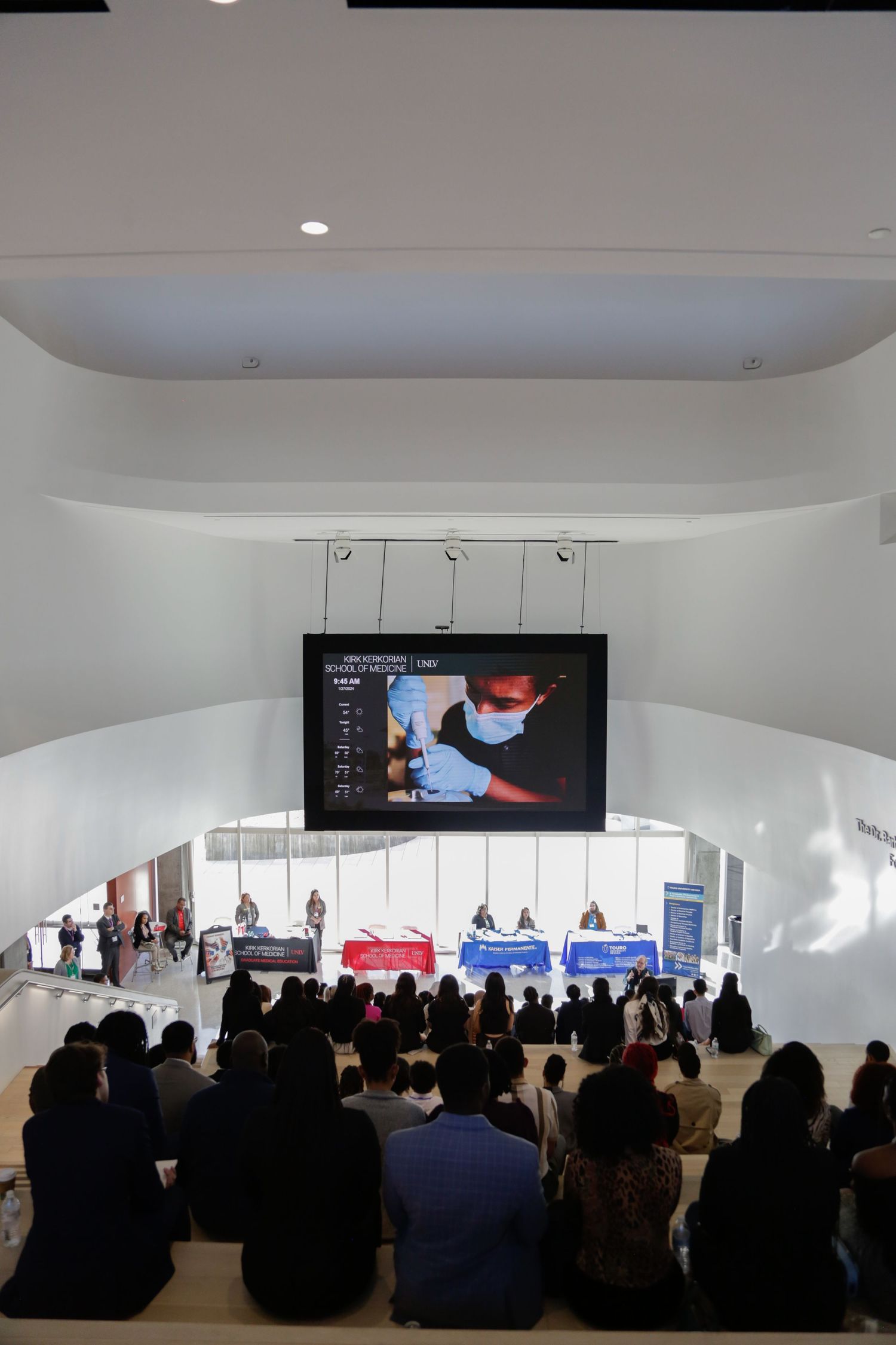 Large audience seated in modern auditorium with curved white walls and digital displays.