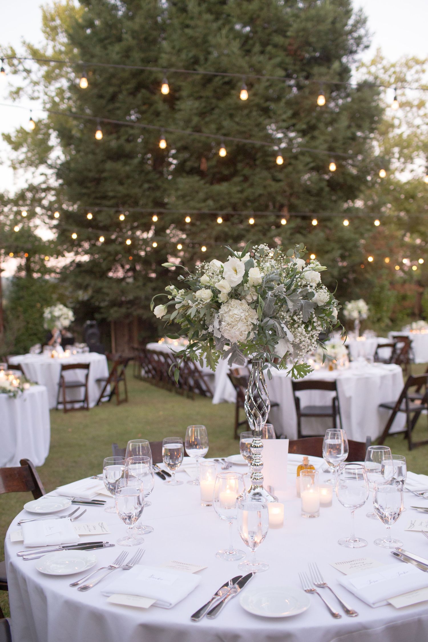 Elegant outdoor wedding reception table with white floral centerpiece and string lights overhead.