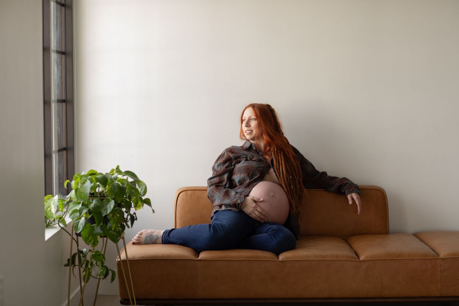 Pregnant woman with long hair relaxing on brown couch with houseplant nearby.