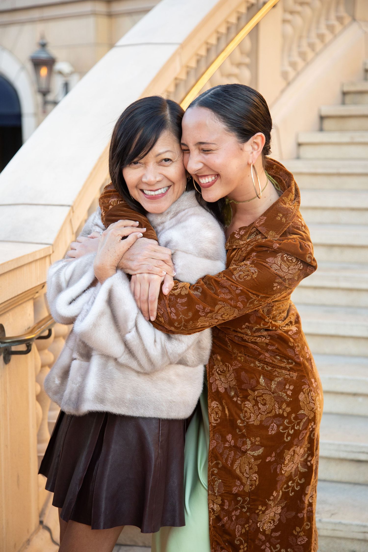Two friends sharing joyful embrace on elegant staircase wearing winter coats.