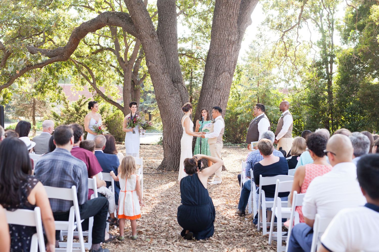 A panoramic view of an outdoor wedding ceremony with white chairs set up under shady trees in a natural setting.